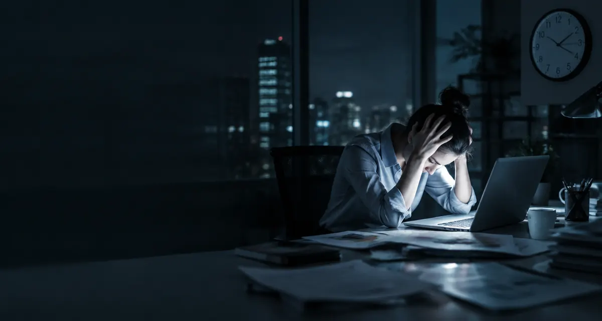 Profissional esgotada com as mãos na cabeça diante do notebook à noite no escritório, ilustrando o Burnout no trabalho